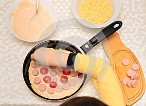 Child hand placing the cheese onto the raw dough for homemade pizza with sausages and tomatoes. Other ingredients are on