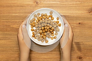 Child hands holding full bowl of yogurt with cereal breakfast  balls on wooden table, top view. Quick and healthy food