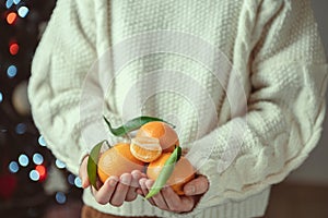 Child hands in knitted sweater holding fresh mandarins with green leaves with bokeh on background