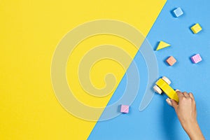 Child hands playing with wooden toy car and colorful building blocks on blue and yellow background, top view