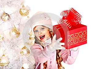 Child in hat and mittens holding red gift box near white Christmas tree.