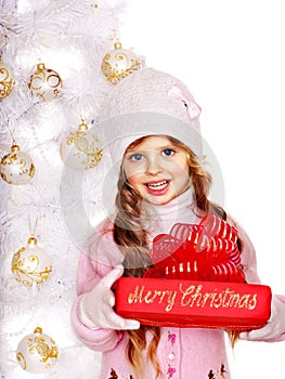 Child in hat and mittens holding red gift box near white Christmas tree.