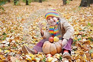 Child having fun with apple and pumpkin in forest, sit on autumn leaves background, fall season