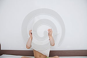 Child hiding behind pillow sitting on bed