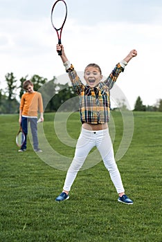 Child holding badminton racquet and triumphing while sibling standing behind