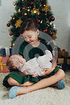 Child holding cute little sisiter while sitting near Christmas tree and gift boxes