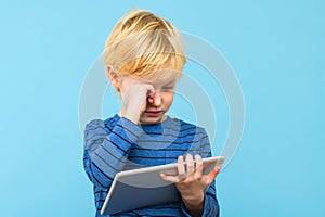 Child holding digital tablet, watching videos, rubbing his eyes, isolated over pastel blue background. Children and technology.