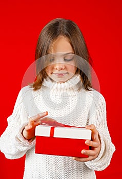 Child holding gift box with red ribbon and looking inside with curiosity on red background