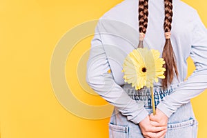 Child holding yellow flower behind her back against yellow background