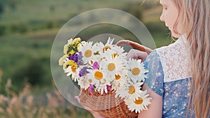 A child holds a basket of flowers, stands in a picturesque place against the backdrop of meadows and green hills
