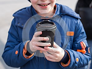 A child holds a black paper cup of drink. Teenage boy holds in hands cup of coffee in paper glass in cold weather.