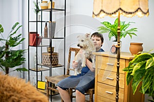 A child holds a dog in his arms and sits on a chair against the wall.