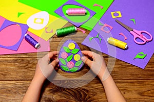 Child holds a felt Easter egg decor in his hands. Child made simple Easter crafts. Sewing tools and materials on a wooden table