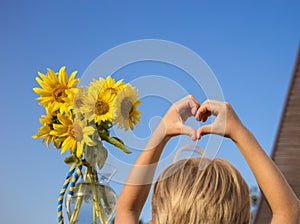child holds hands in shape of heart against background of sky and blooming sunflowers