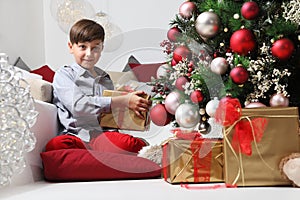 Child holds in his hands the Christmas gift box near the tree decorated with balls, in living room sitting on the floor near the