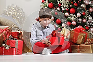 Child holds in his hands the Christmas gift box near the tree decorated with balls, in living room sitting on the floor near the