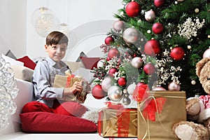 Child holds in his hands the Christmas gift box near the tree decorated with balls, in living room sitting on the floor near the