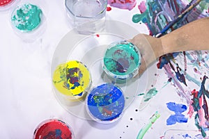 The child holds a jar of watercolor paint in his hand against the background of his drawings. Kids creativity