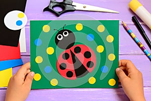 Child holds a summer card in his hands. Child made a paper ladybug card. Stationery on a wooden table. Easy paper circle crafts