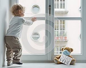 Child in home quarantine playing at the window with his sick teddy bear wearing a medical mask against viruses during coronavirus