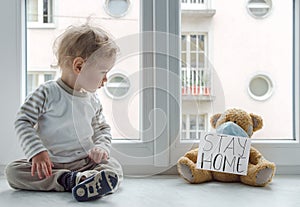 Child in home quarantine playing at the window with his sick teddy bear wearing a medical mask against viruses during coronavirus