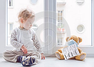 Child in home quarantine playing at the window with his sick teddy bear wearing a medical mask against viruses during coronavirus