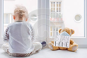 Child in home quarantine playing at the window with his sick teddy bear wearing a medical mask against viruses during coronavirus