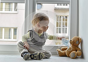 Child in home quarantine at the window putting a medical mask on his sick teddy bear, for protection against viruses during