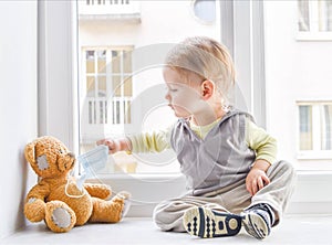 Child in home quarantine at the window putting a medical mask on his sick teddy bear, for protection against viruses during