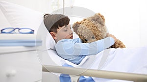Child in hospital lying alone in bed play with teddy bear hugs him and smiling in camera