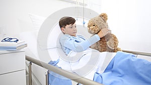 Child in hospital lying alone in bed play with teddy bear hugs him and smiling in camera