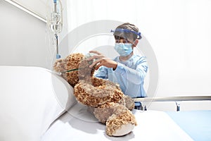 Child in the hospital puts oxygen mask on teddy bear on bed, wearing protective visor and surgical mask, corona virus covid 19