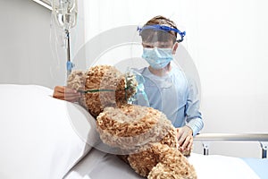 Child in the hospital puts oxygen mask on teddy bear on bed, wearing protective visor and surgical mask, corona virus covid 19