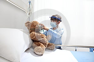 Child in the hospital puts oxygen mask on teddy bear on bed, wearing protective visor and surgical mask, corona virus covid 19