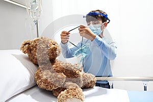 Child in the hospital puts oxygen mask on teddy bear on bed, wearing protective visor and surgical mask, corona virus covid 19