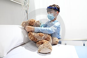 Child in the hospital puts oxygen mask on teddy bear on bed, wearing protective visor and surgical mask, corona virus covid 19