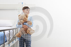 Child in hospital room standing next to bed hugging a teddy bear wearing a pajama isolated on white wall