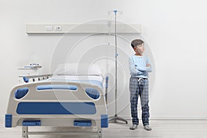 Child in hospital room standing next to bed wearing a pajama isolated on white wall
