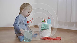 Child with household chemicals sitting near a bucket with plastic bottles, kid boy sprays detergent on rubber glove and