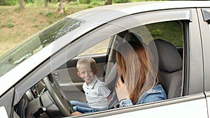 A child indulges in a car sitting in the front seat, mother talking on the phone