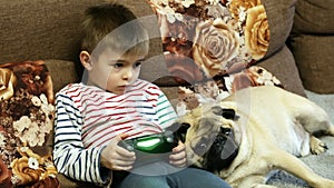 Child with a joystick in his hands playing computer games, pug dog with child on the couch at home