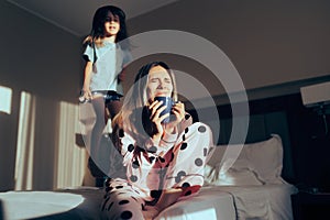 Child Jumping in Bed Next to Exasperated Mom Drinking Coffee