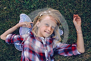 child kid girl with long hair wearing pink fairy wings and plaid shirt, lying on grass