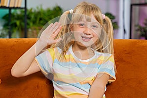 Child kid girl smiling friendly at camera, waving hands gesturing hello hi greeting at home on couch