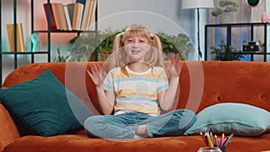 Child kid girl smiling friendly at camera, waving hands gesturing hello hi greeting at home on couch