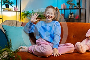 Child kid girl smiling friendly at camera, waving hands gesturing hello hi greeting at home on couch