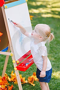 Child kid girl standing outside in summer autumn park drawing on easel with markers looking away playing studying