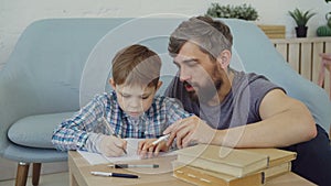 Child is learning, writing words in exercise book and his father is teaching him sitting near table and explaining rules