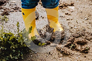 Child legs in yellow muddy rubber boots on wet mud.  Baby playing with dirt at rainy weather.