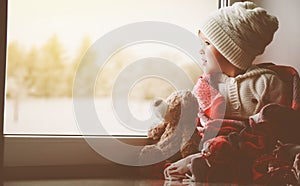 Child little girl with teddy bear at window and looking at wint